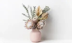 Un bouquet di fiori secchi, tra cui protee e foglie di palma dorate, in un vaso rosa. Questi fiori statici non hanno bisogno di acqua per mantenere la loro bellezza.