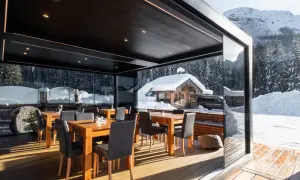 Tavoli e sedie sotto una moderna pergola bioclimatica, con vista su un paesaggio invernale innevato e un rifugio alpino.