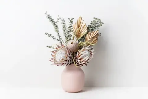 Un bouquet di fiori secchi, tra cui protee e foglie di palma dorate, in un vaso rosa. Questi fiori statici non hanno bisogno di acqua per mantenere la loro bellezza.