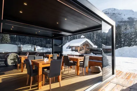 Tavoli e sedie sotto una moderna pergola bioclimatica, con vista su un paesaggio invernale innevato e un rifugio alpino.