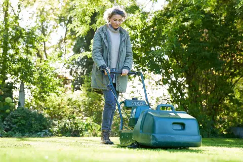 Donna con tosaerba verde, impegnata a tagliare erba bagnata in un giardino soleggiato.