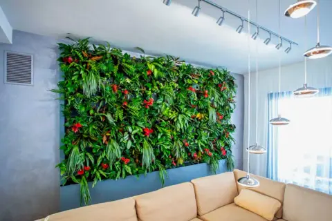 Un lussureggiante giardino verticale interno, con piante da parete verticale rigogliose e fiori rossi, domina la stanza sopra un divano beige.