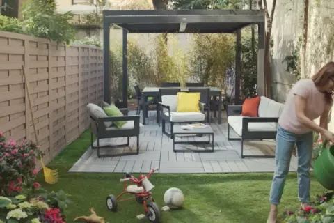 Donna annaffia fiori in un giardino moderno con mobili da esterno e un pergolato. Giardini idee da copiare per un'oasi urbana.