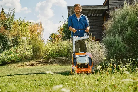 Uomo con tosaerba elettrico su un prato verde, con fiori e vegetazione rigogliosa. Un perfetto prato primavera.