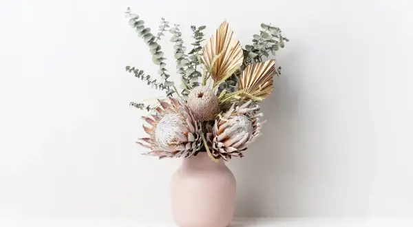Un bouquet di fiori secchi, tra cui protee e foglie di palma dorate, in un vaso rosa. Questi fiori statici non hanno bisogno di acqua per mantenere la loro bellezza.