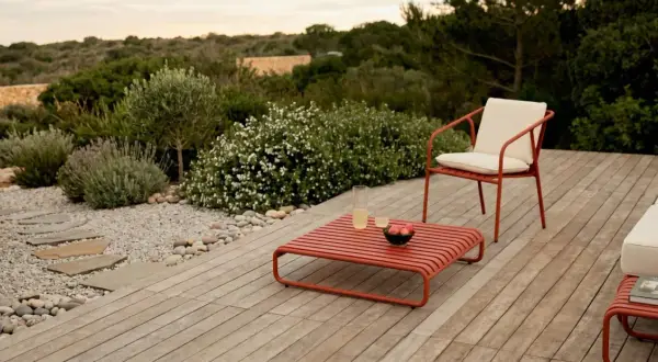 Un elegante patio in legno con mobili rossi, un sentiero di ghiaia e piante mediterranee. Perfetto per idee giardino senza prato.