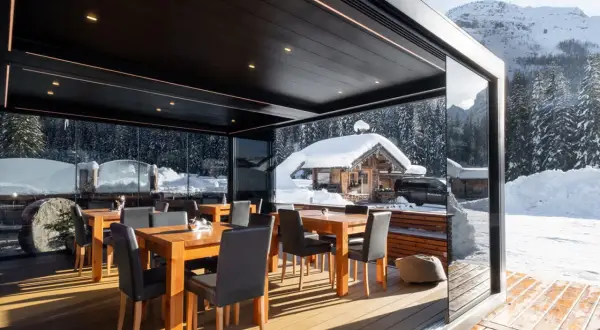 Tavoli e sedie sotto una moderna pergola bioclimatica, con vista su un paesaggio invernale innevato e un rifugio alpino.