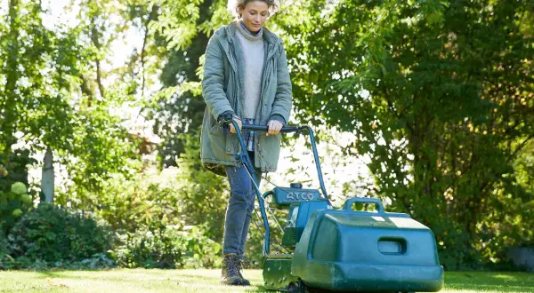 Donna con tosaerba verde, impegnata a tagliare erba bagnata in un giardino soleggiato.