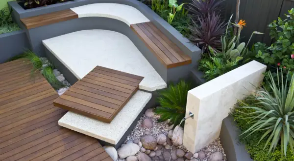 Un elegante giardino moderno con sedute in legno, pavimentazione in ghiaia e una piccola fontana.