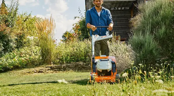 Uomo con tosaerba elettrico su un prato verde, con fiori e vegetazione rigogliosa. Un perfetto prato primavera.