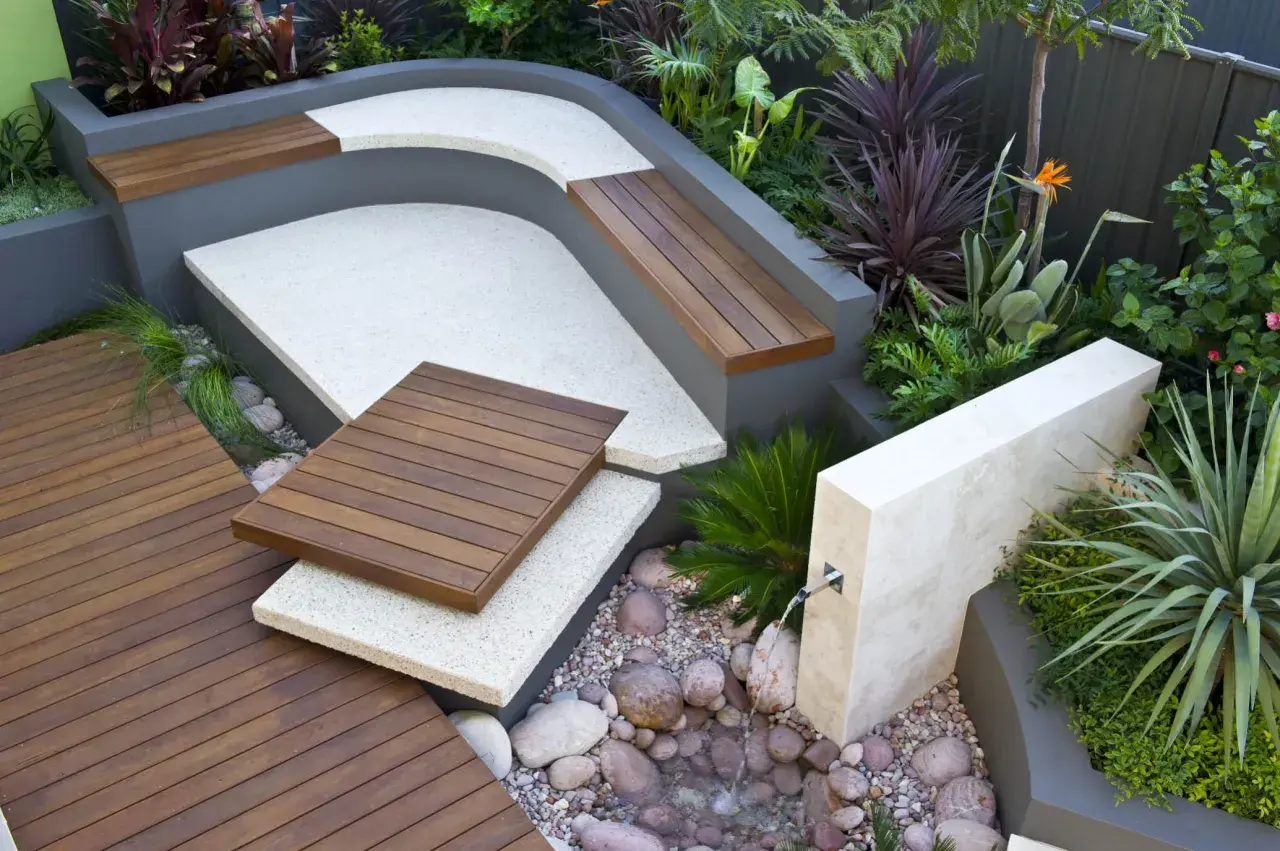 Un elegante giardino moderno con sedute in legno, pavimentazione in ghiaia e una piccola fontana.