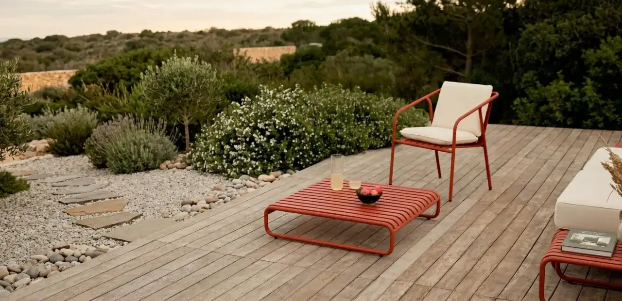 Un elegante patio in legno con mobili rossi, un sentiero di ghiaia e piante mediterranee. Perfetto per idee giardino senza prato.