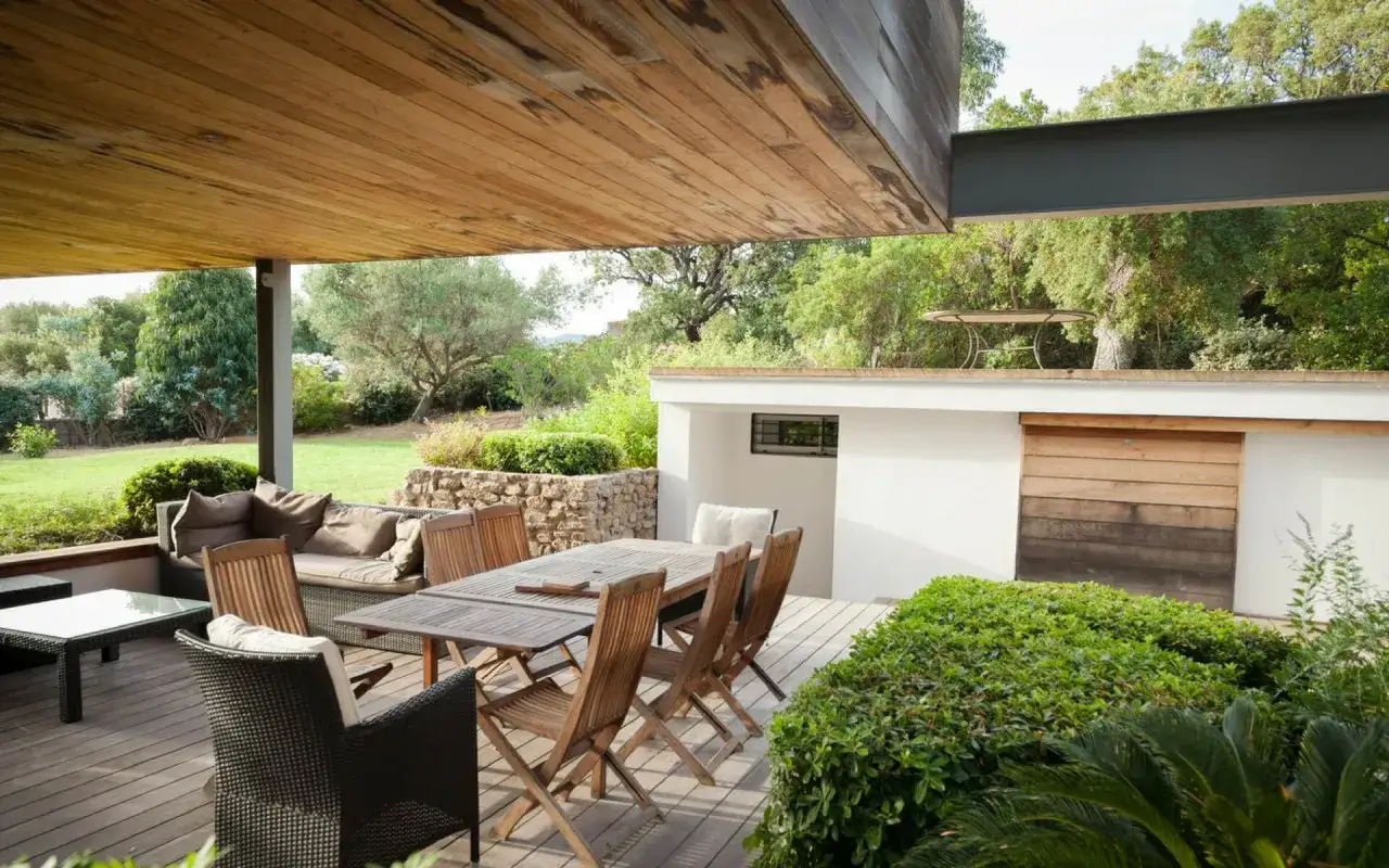 Arredamento esterno: tavolo, sedie e divano su un deck in legno, con un piccolo giardino davanti casa.