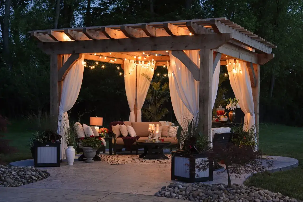 Pergolato illuminato con luci soffuse, tende bianche e arredi da esterno, creando piccoli giardini da sogno.