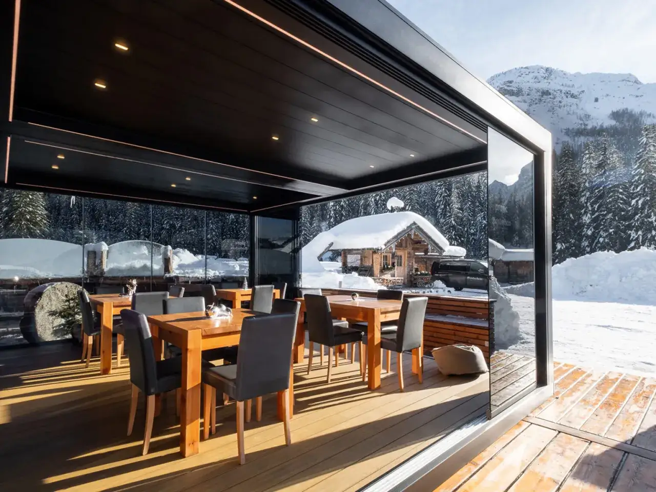 Tavoli e sedie sotto una moderna pergola bioclimatica, con vista su un paesaggio invernale innevato e un rifugio alpino.