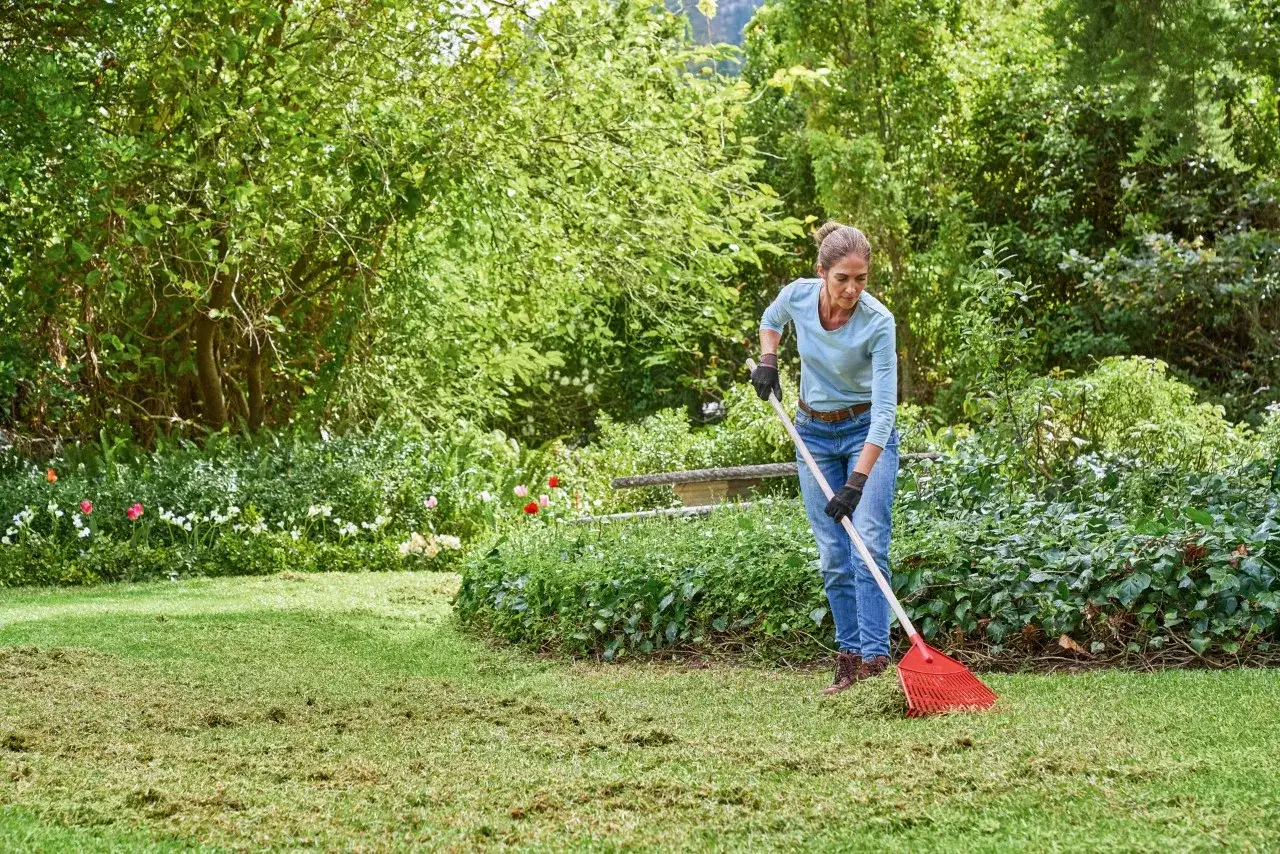 Donna rastrella il prato in primavera, raccogliendo l'erba tagliata tra fiori colorati e alberi rigogliosi.
