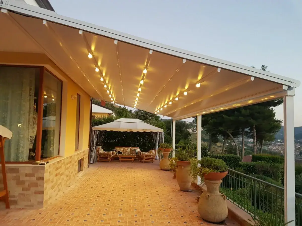 Tettoia addossata con luci soffuse illumina un patio con arredi da esterno e vasi decorativi, creando un'atmosfera accogliente.