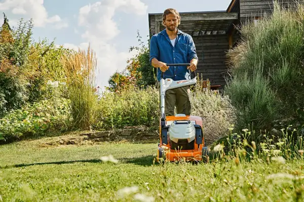 Uomo con tosaerba elettrico su un prato verde, con fiori e vegetazione rigogliosa. Un perfetto prato primavera.
